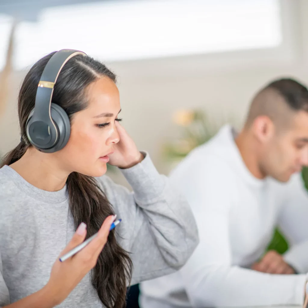 Person wearing headphones adjusting them while working at a desk, with another person working in the background.