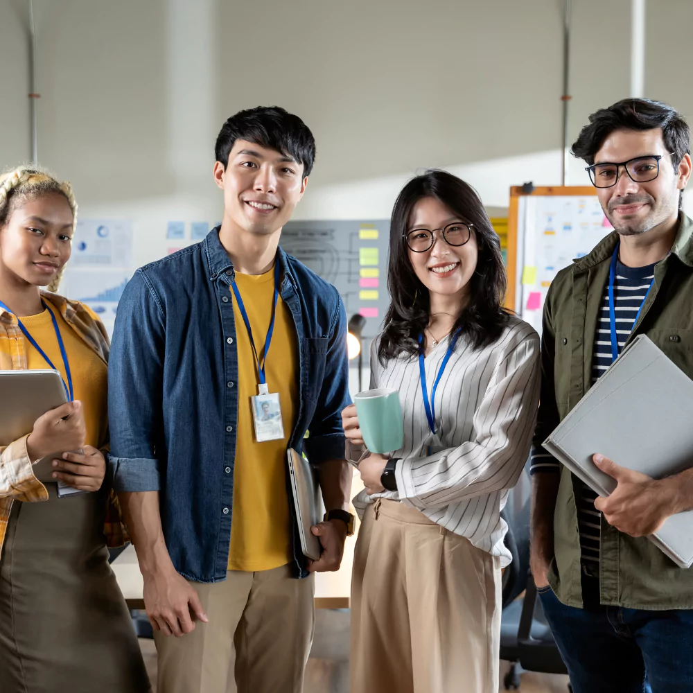A team of four colleagues standing together in a bright office, smiling at the camera and holding work materials.