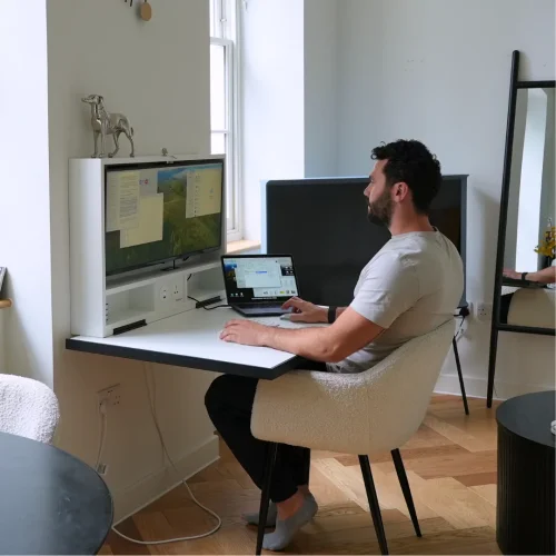 Person working at an open DropTop desk with monitor and laptop by a window.
