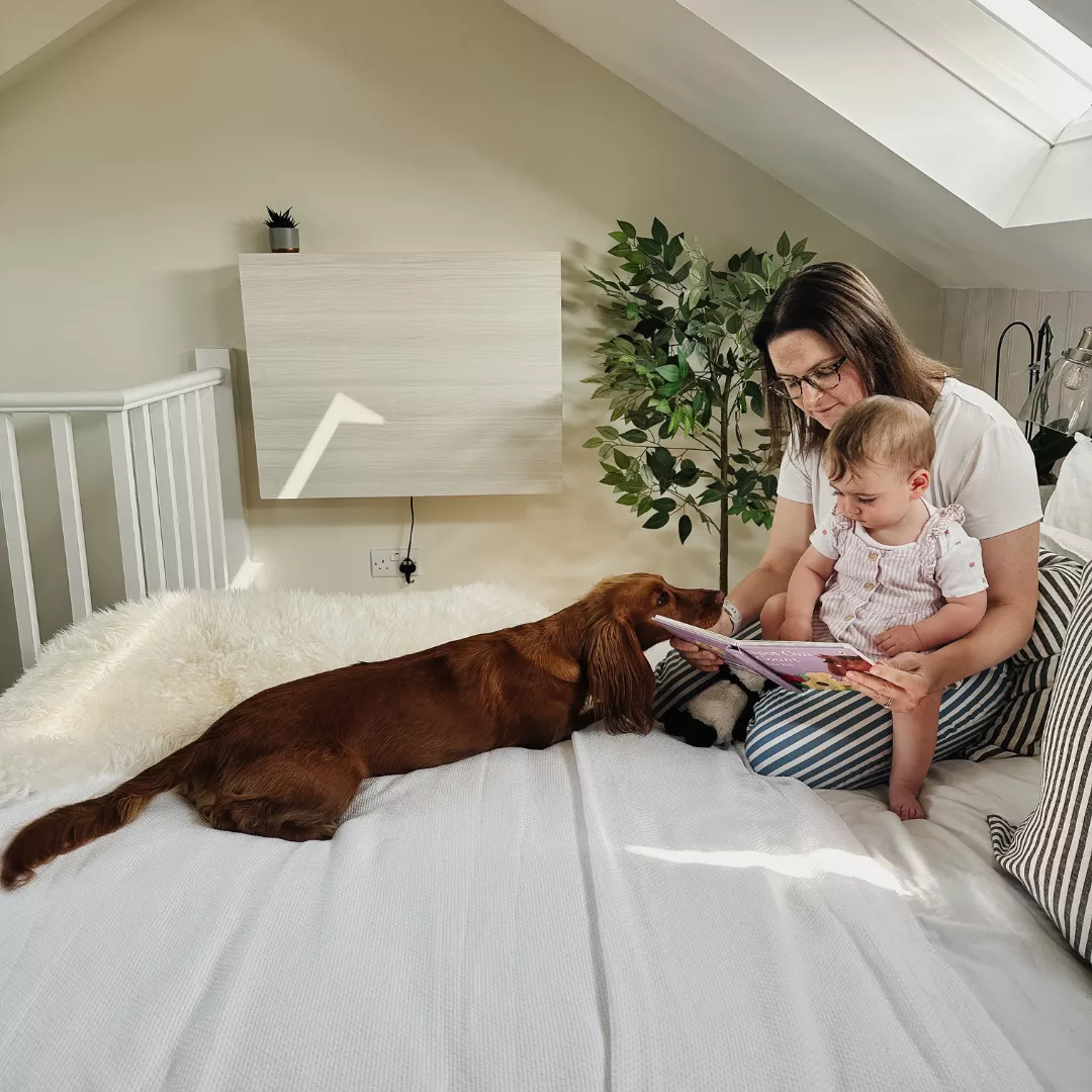 Person sitting on a bed reading with a child and dog in a bedroom, with a closed DropTop desk mounted on the wall in the background.