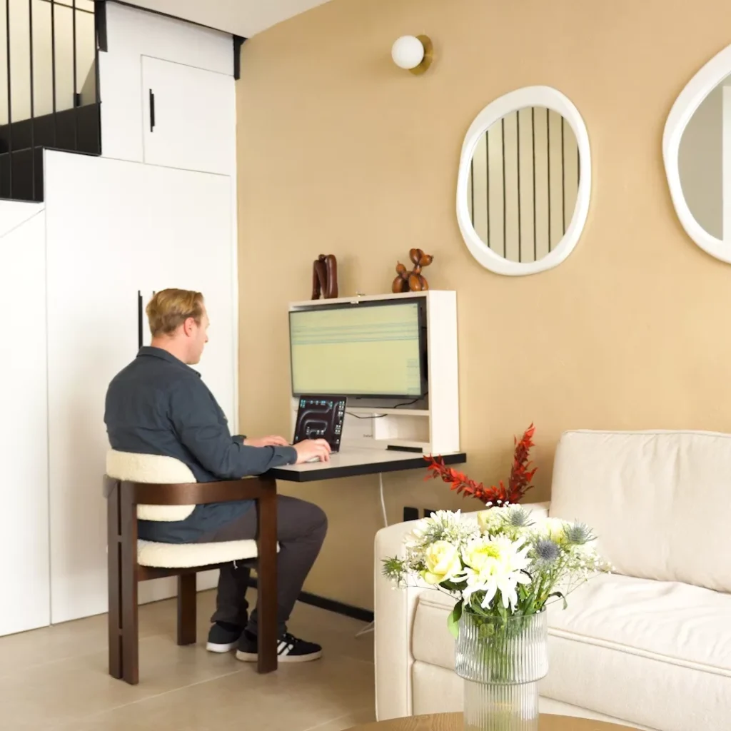 Person working at an open DropTop desk with monitor and laptop in a living room.